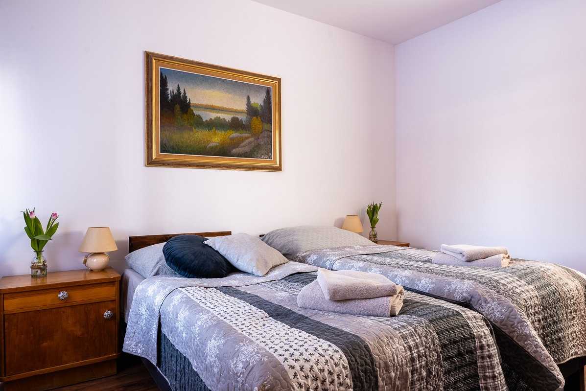 Jasny pokój gościnny z dwoma łóżkami, nad którymi wisi obraz w złotej ramie. Na szafce nocnej stoi lampka i wazon z tulipanami. Przytulne wnętrze.