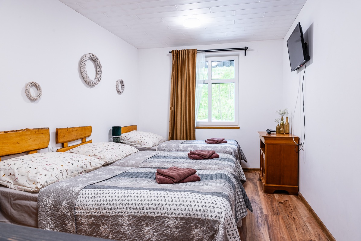 Jasny pokój gościnny z trzema łóżkami, drewnianymi zagłówkami i dekoracjami ściennymi w postaci wianków. Na komodzie bukiet w butelkach. Telewizor na ścianie.