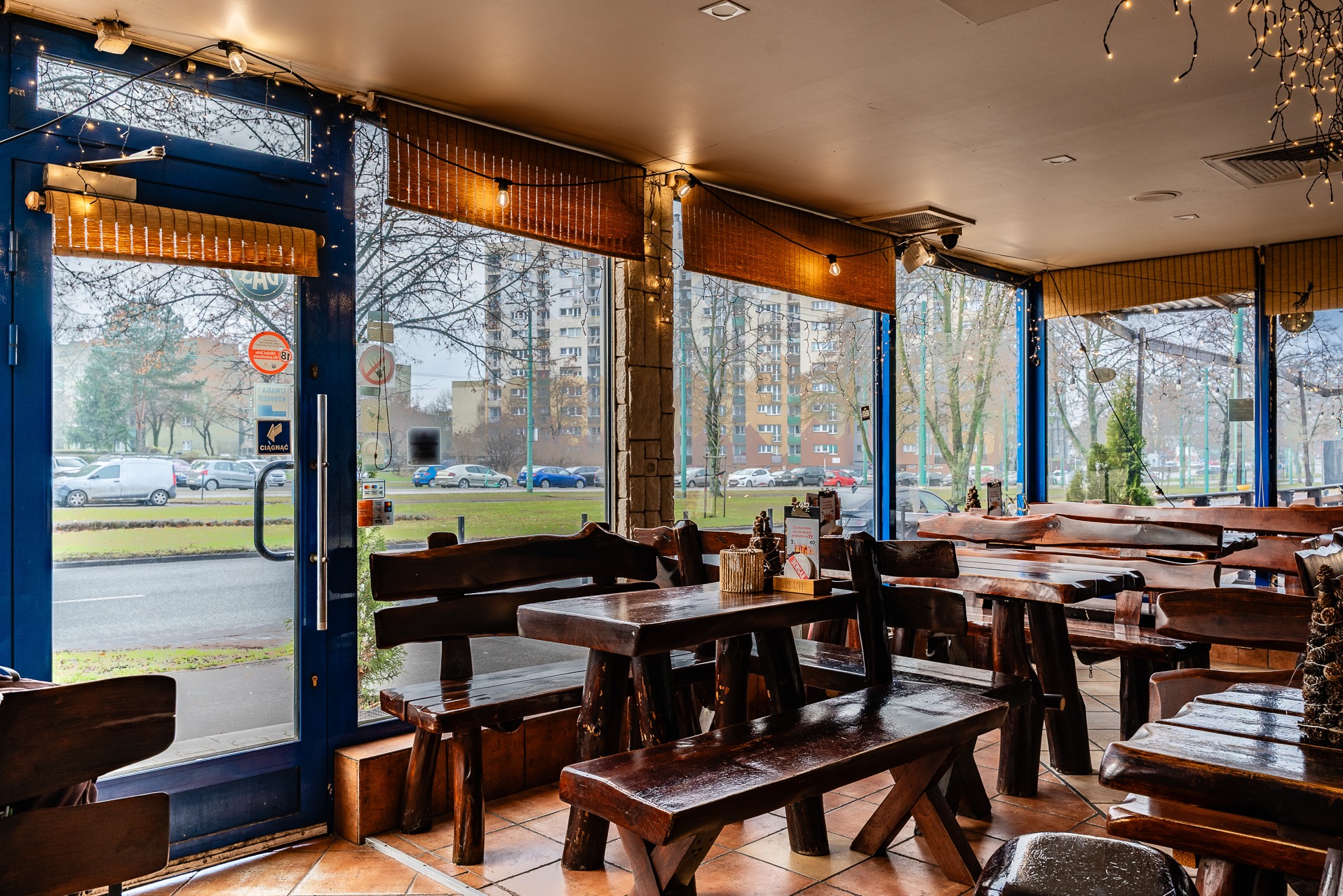 Wnętrze baru z drewnianymi stołami i ławami w rustykalnym stylu, widok na ulicę przez duże okna z roletami bambusowymi i dekoracyjnymi lampkami.