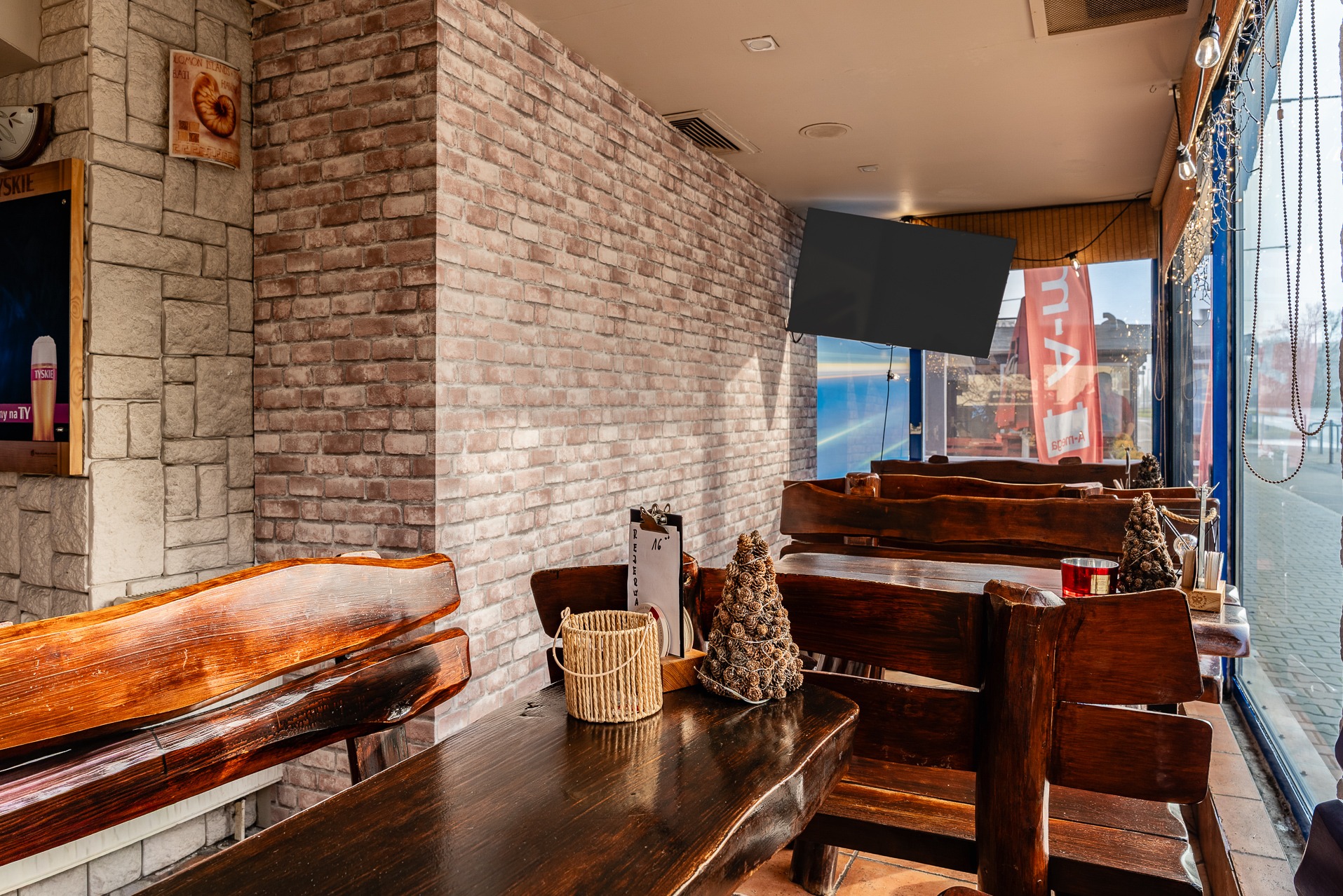 Rustykalne wnętrze baru z drewnianymi stołami i krzesłami, ceglaną ścianą i dekoracjami. Widok na ulicę przez okno. Klimatyczne oświetlenie.