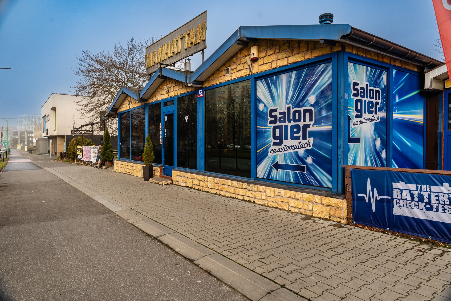 Zewnętrzny widok budynku z napisem 'Salon gier na automatach' na oknach i szyldem 'Manhattan' na dachu. Żółta cegła i niebieskie ramy okienne.