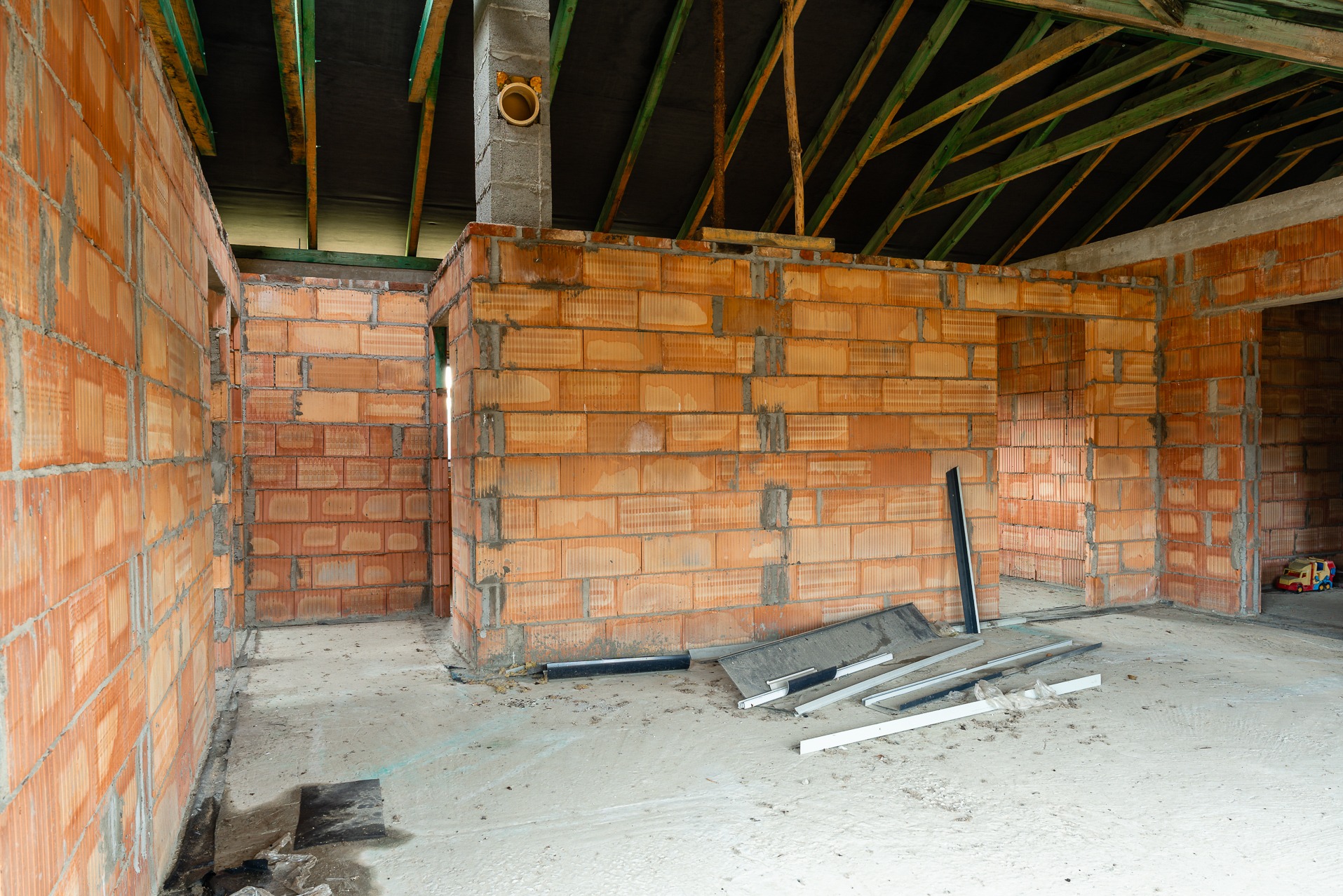 Wnętrze domu w stanie surowym otwartym: pomarańczowe ściany z pustaków, surowy beton na podłodze, drewniana konstrukcja dachu. Widoczne narzędzia budowlane.