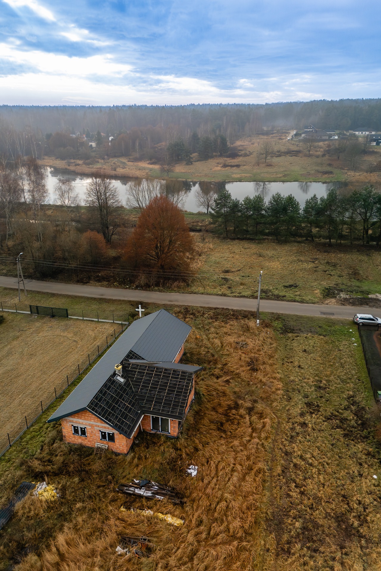Zdjęcie z drona: dom w stanie surowym otwartym z częściowo położonym dachem na tle rzeki i lasu. Widoczna droga, samochód i krzyż. Sesja dla agencji nieruchomości.