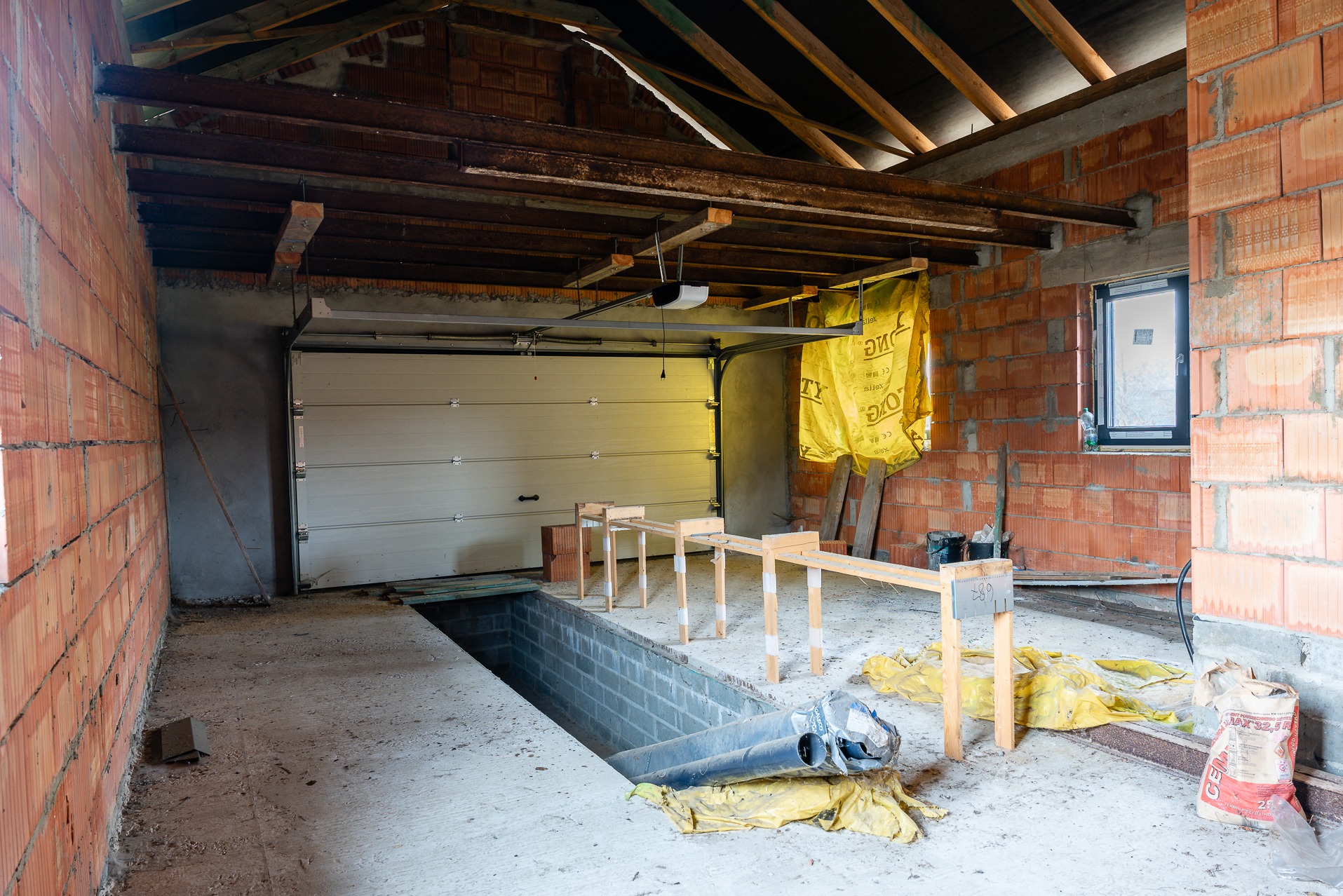 Wnętrze surowego garażu z otwartą bramą, widoczne ceglane ściany, drewniane belki stropowe, kanał samochodowy i materiały budowlane. Stan deweloperski.