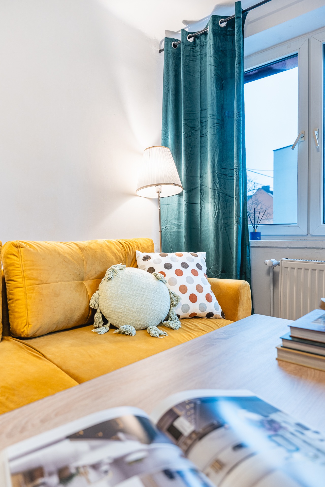 Jasny salon z żółtą sofą, dekoracyjnymi poduszkami, lampą podłogową i otwartą książką na stole. W tle okno z turkusową zasłoną i grzejnik.