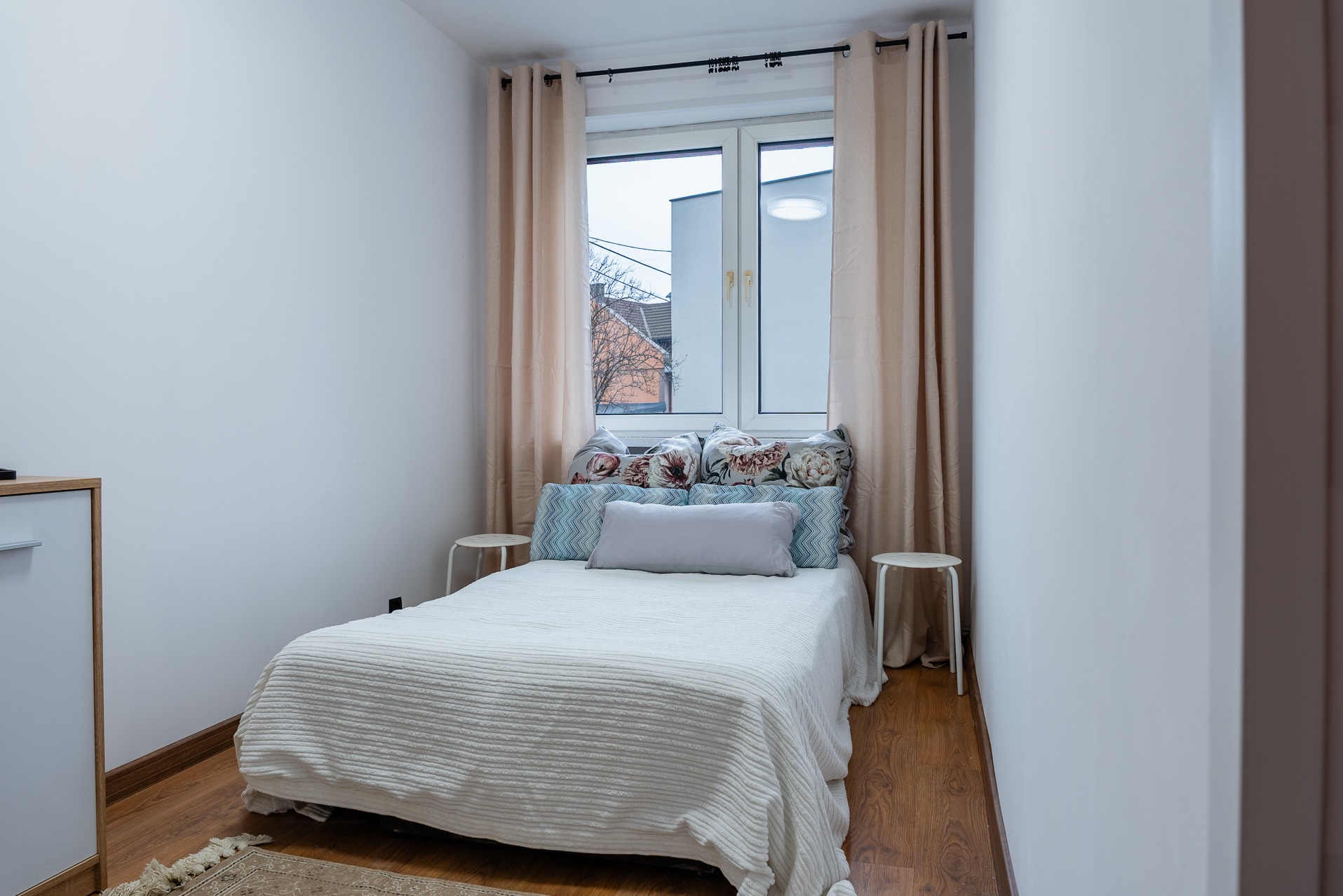 Jasna sypialnia z łóżkiem, wzorzystymi poduszkami i beżowymi zasłonami. Widok z okna na budynki. Minimalistyczny wystrój, drewniana podłoga i dywanik.