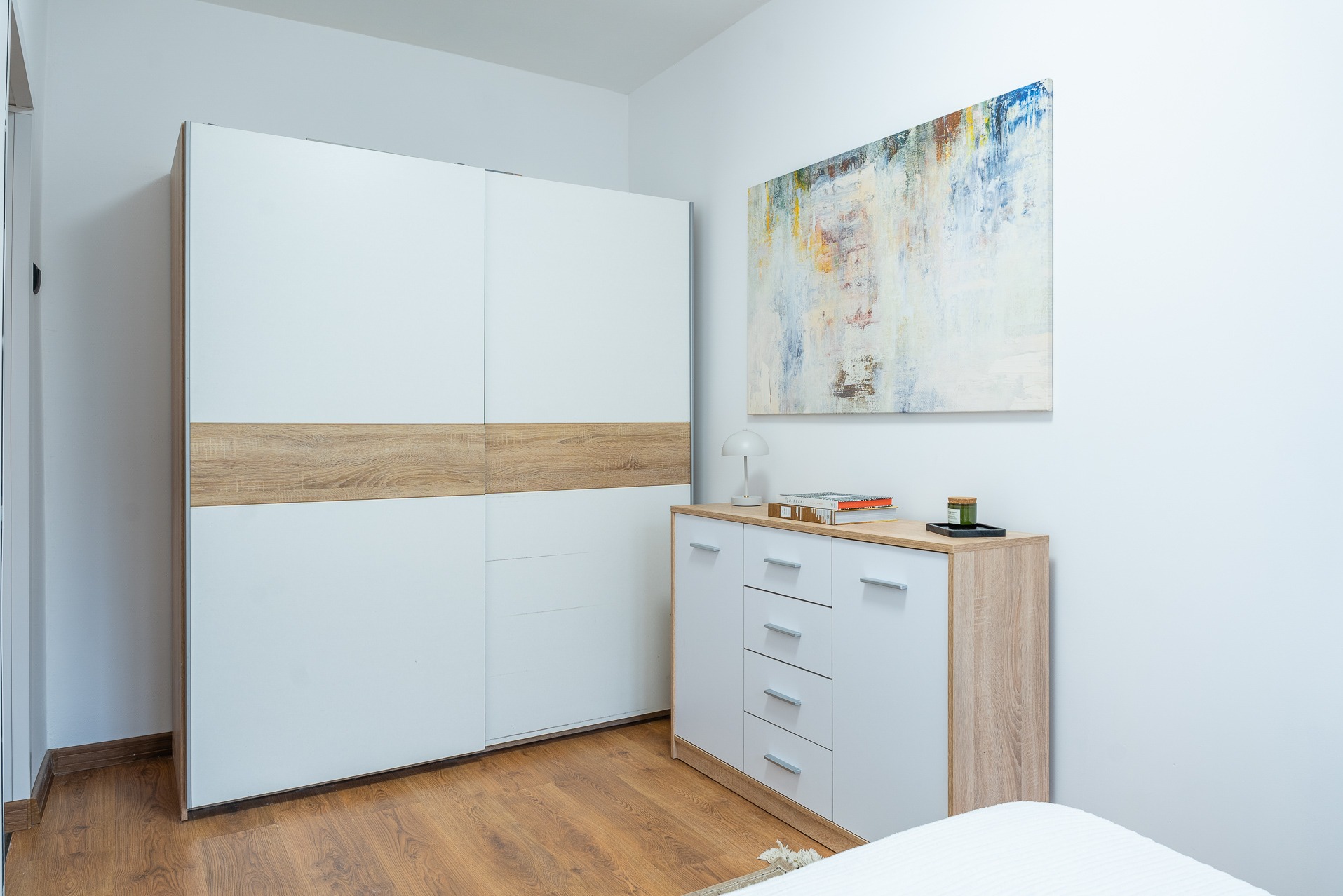 Jasne wnętrze sypialni: biała szafa przesuwna z drewnianym akcentem, komoda z lampką i abstrakcyjny obraz na ścianie. Minimalistyczny design, neutralna kolorystyka.