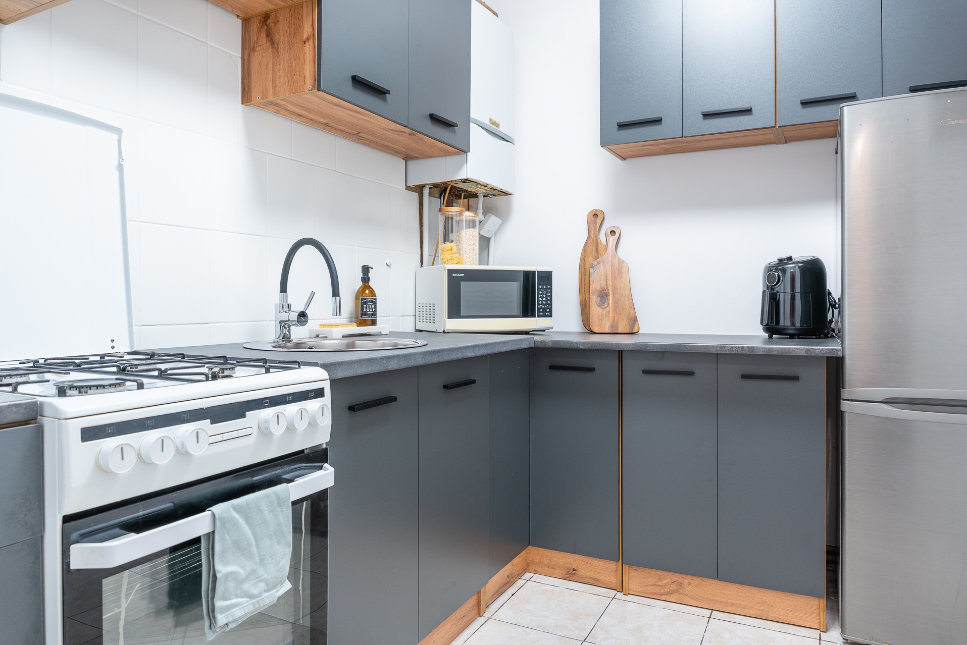 Funkcjonalna kuchnia w szarościach: zabudowa z drewnianymi akcentami, sprzęt AGD (lodówka, kuchenka, mikrofalówka), deski do krojenia i armatura. Jasne płytki na ścianie.