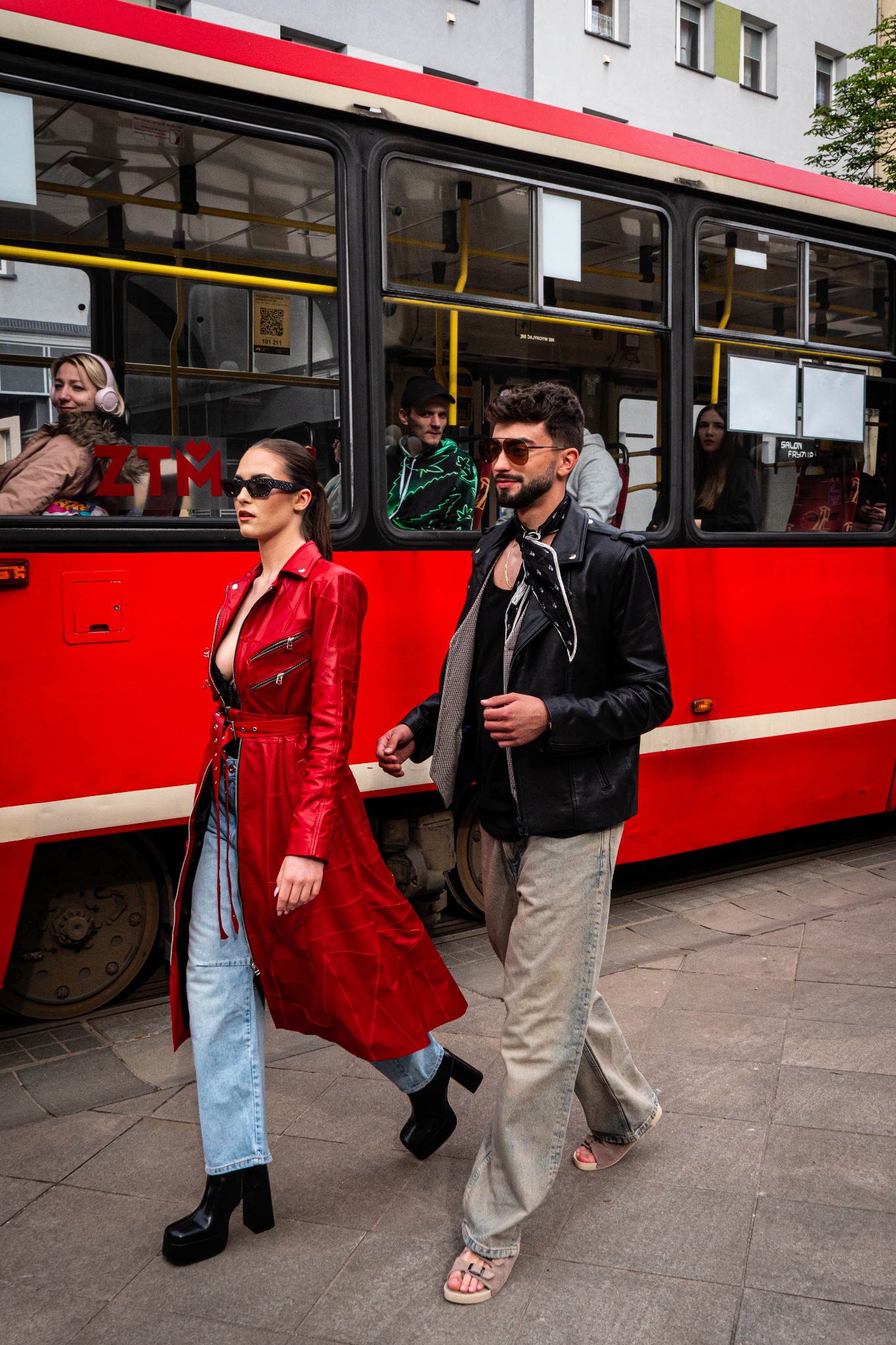 Stylowa sesja: kobieta w czerwonym płaszczu i mężczyzna w skórzanej kurtce spacerują na tle czerwonego tramwaju miejskiego. Uliczny szyk, moda miejska.