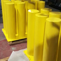 Seria jaskrawożółtych słupków zabezpieczających, świeżo po nałożeniu farby, ustawionych na palecie w hali produkcyjnej.
