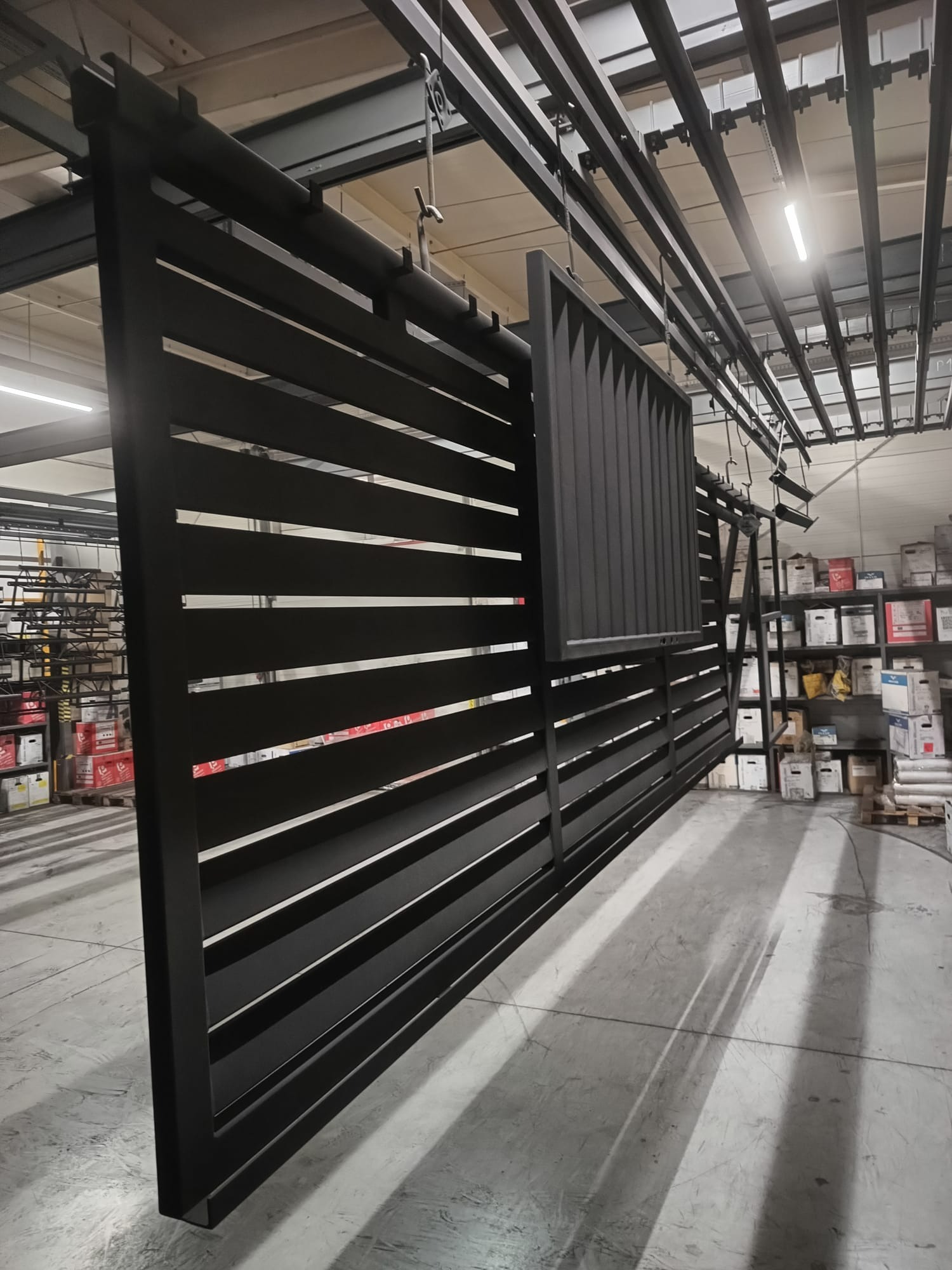 Części metalowego ogrodzenia, zawieszone na szynach, przygotowane do dalszej obróbki lub transportu w hali produkcyjnej.