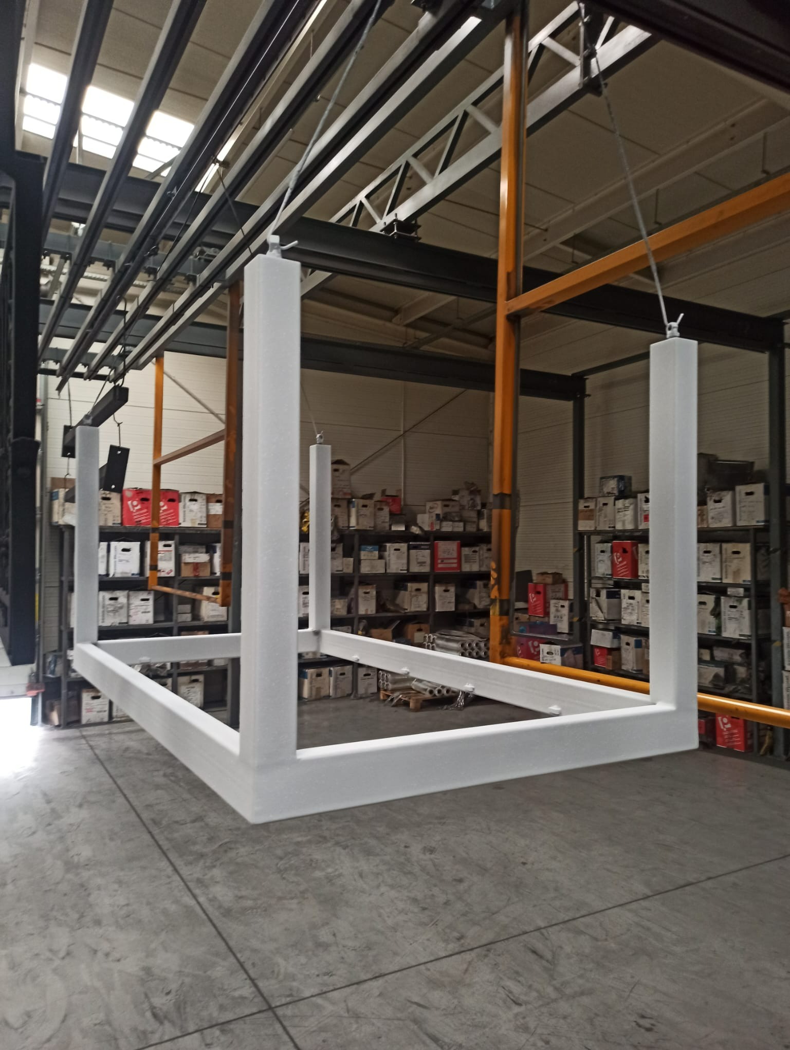 Metalowa konstrukcja w kształcie ramy, świeżo po malowaniu na biało, zawieszona na hakach w hali produkcyjnej.