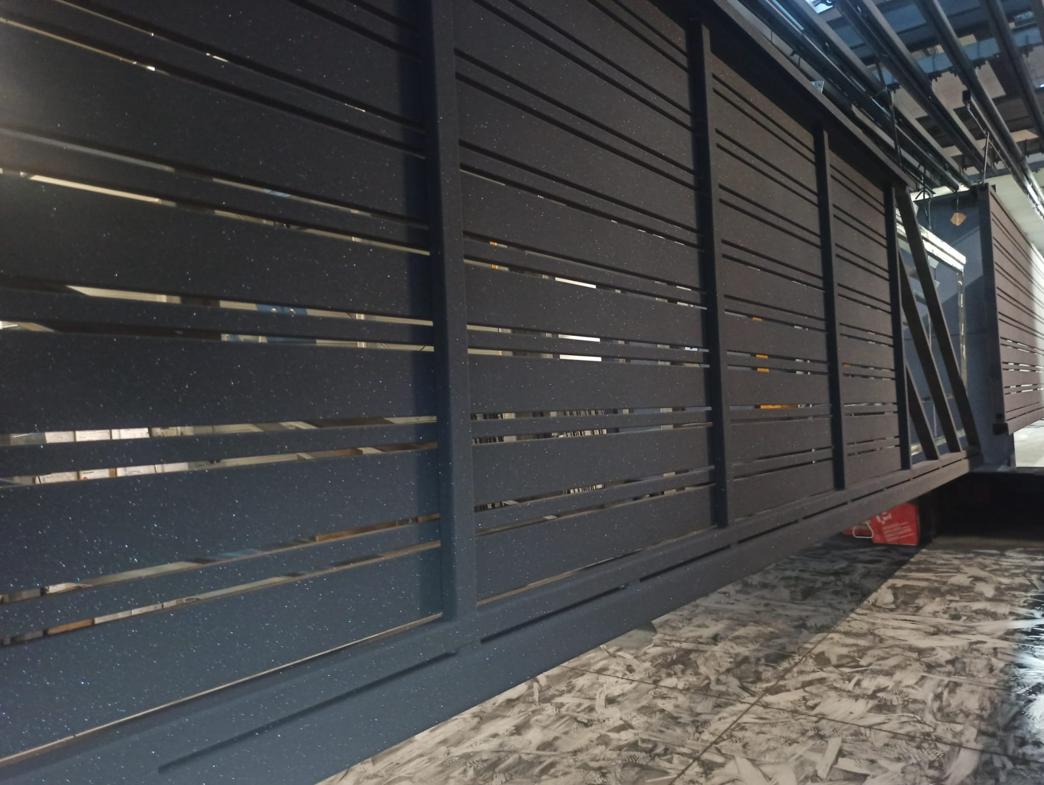 Ciemnoszara metalowa brama przesuwna z poziomymi szczelinami, wykończona farbą proszkową z efektem brokatu, widoczna z perspektywy pod kątem na tle wzorzystej podłogi.