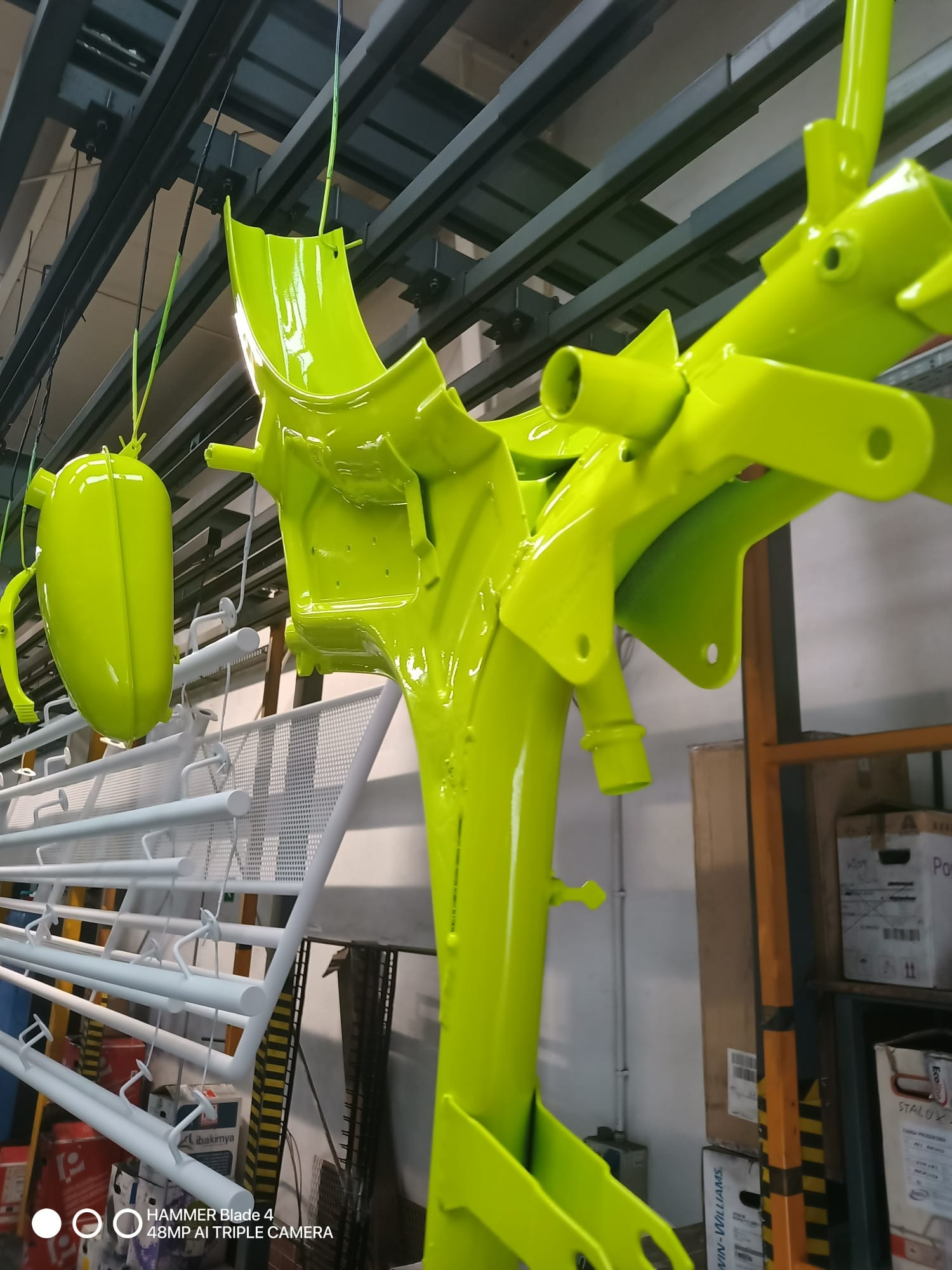 Elementy metalowe motocykla zawieszone na linii produkcyjnej po nałożeniu jaskrawozielonej powłoki. W tle widoczne białe elementy i regał z pudełkami.