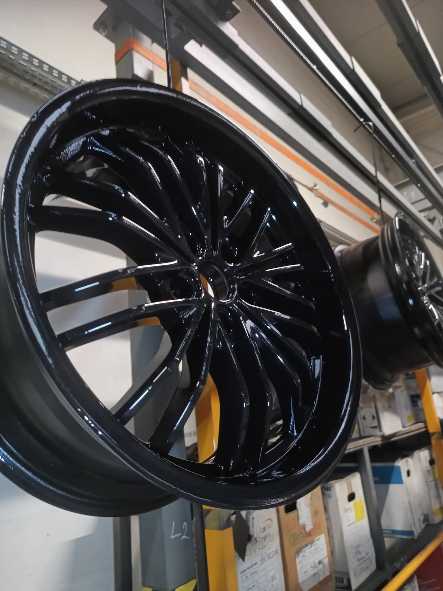 Błyszczące, czarne felgi samochodowe zawieszone na linii produkcyjnej, widoczne ślady po procesie lakierniczym, w tle kartony i elementy konstrukcyjne hali.