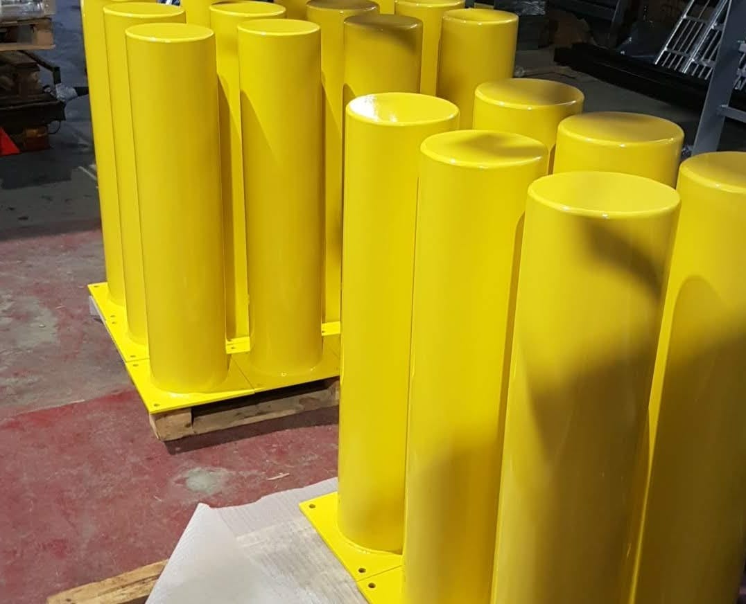 Seria jaskrawożółtych słupków zabezpieczających, świeżo po nałożeniu farby, ustawionych na palecie w hali produkcyjnej.