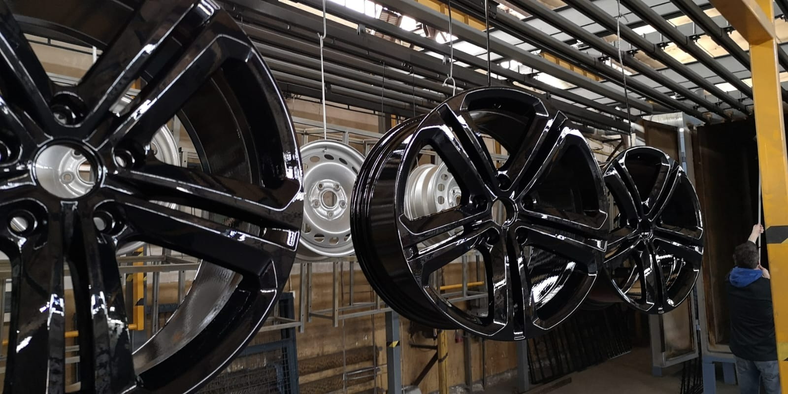 Błyszczące, czarne felgi samochodowe zawieszone na metalowym stelażu w hali produkcyjnej, prawdopodobnie tuż po procesie lakierowania.