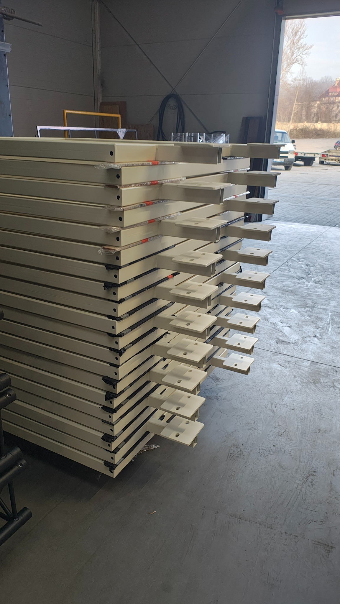 Stos pomalowanych na beżowo metalowych elementów ogrodzeniowych z przyspawanymi uchwytami, przygotowanych do transportu, widoczne ślady po wózkach widłowych na posadzce hali.
