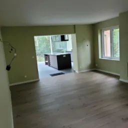 Pomalowane na oliwkowo ściany i ułożone panele podłogowe w nowym, pustym pokoju z widokiem na kuchnię z ciemną zabudową wyspową.