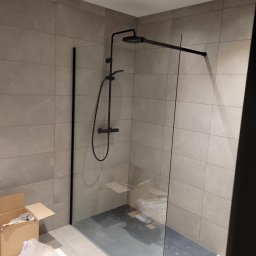 Dominika Kazuch - Nowoczesna kabina prysznicowa typu walk-in z czarną armaturą na tle szarych płytek, widoczne kartony i folia ochronna sugerujące prace wykończeniowe.