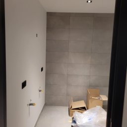 Dominika Kazuch - Łazienka w trakcie remontu: szare płytki na ścianie, widoczne wystające rury z żółtymi zabezpieczeniami, niepodłączone gniazdka elektryczne, kartony i folia na podłodze, widok z korytarza przez...