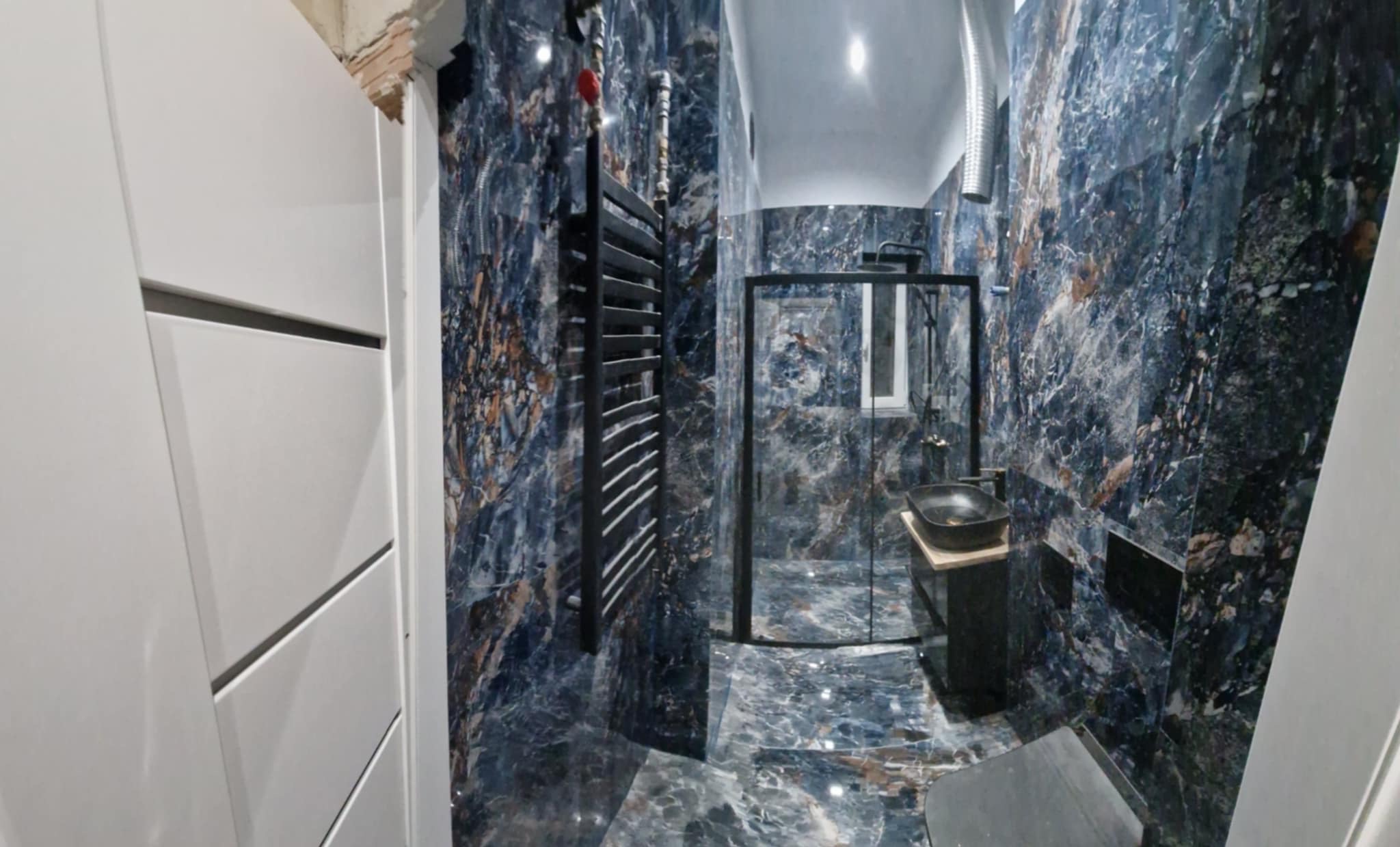 Nowoczesna łazienka z ciemnymi, marmurkowymi płytkami na ścianach i podłodze, czarną armaturą i grzejnikiem drabinkowym, kabiną prysznicową ze szkła i czarną umywalką nablatową.