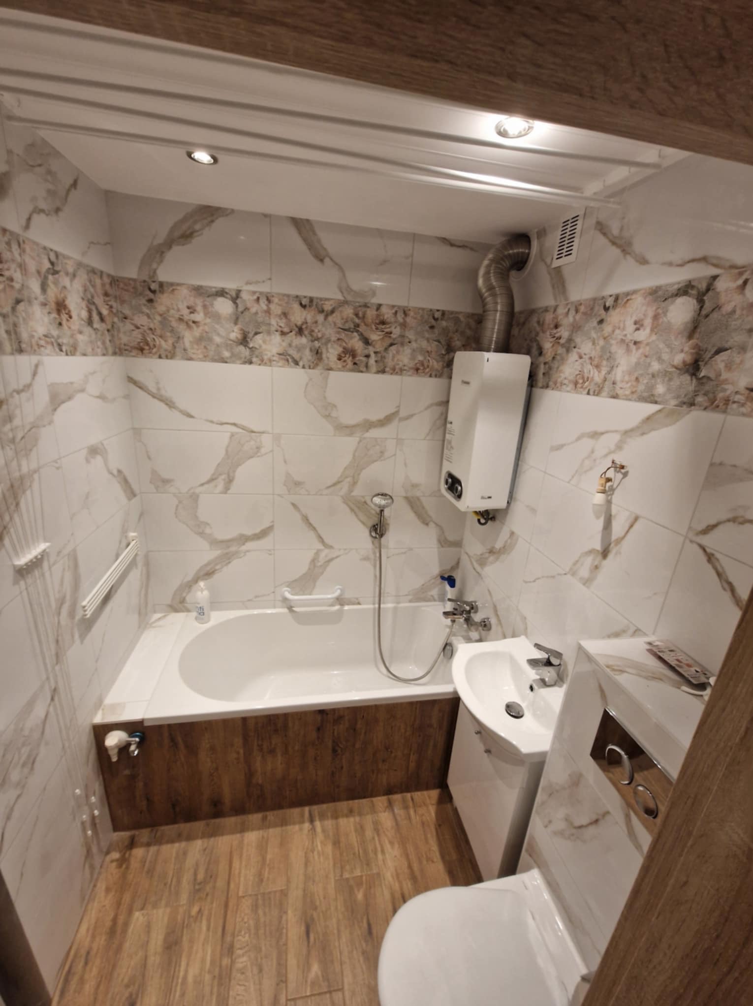 Nowoczesna łazienka z płytkami imitującymi marmur, drewnopodobną podłogą i obudową wanny, oraz białym piecykiem gazowym.