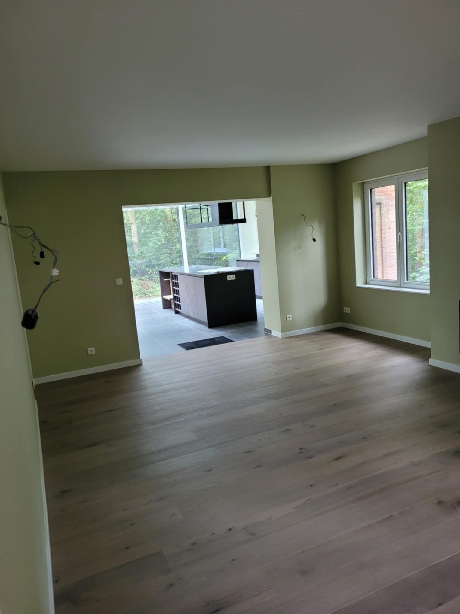 Pomalowane na oliwkowo ściany i ułożone panele podłogowe w nowym, pustym pokoju z widokiem na kuchnię z ciemną zabudową wyspową.