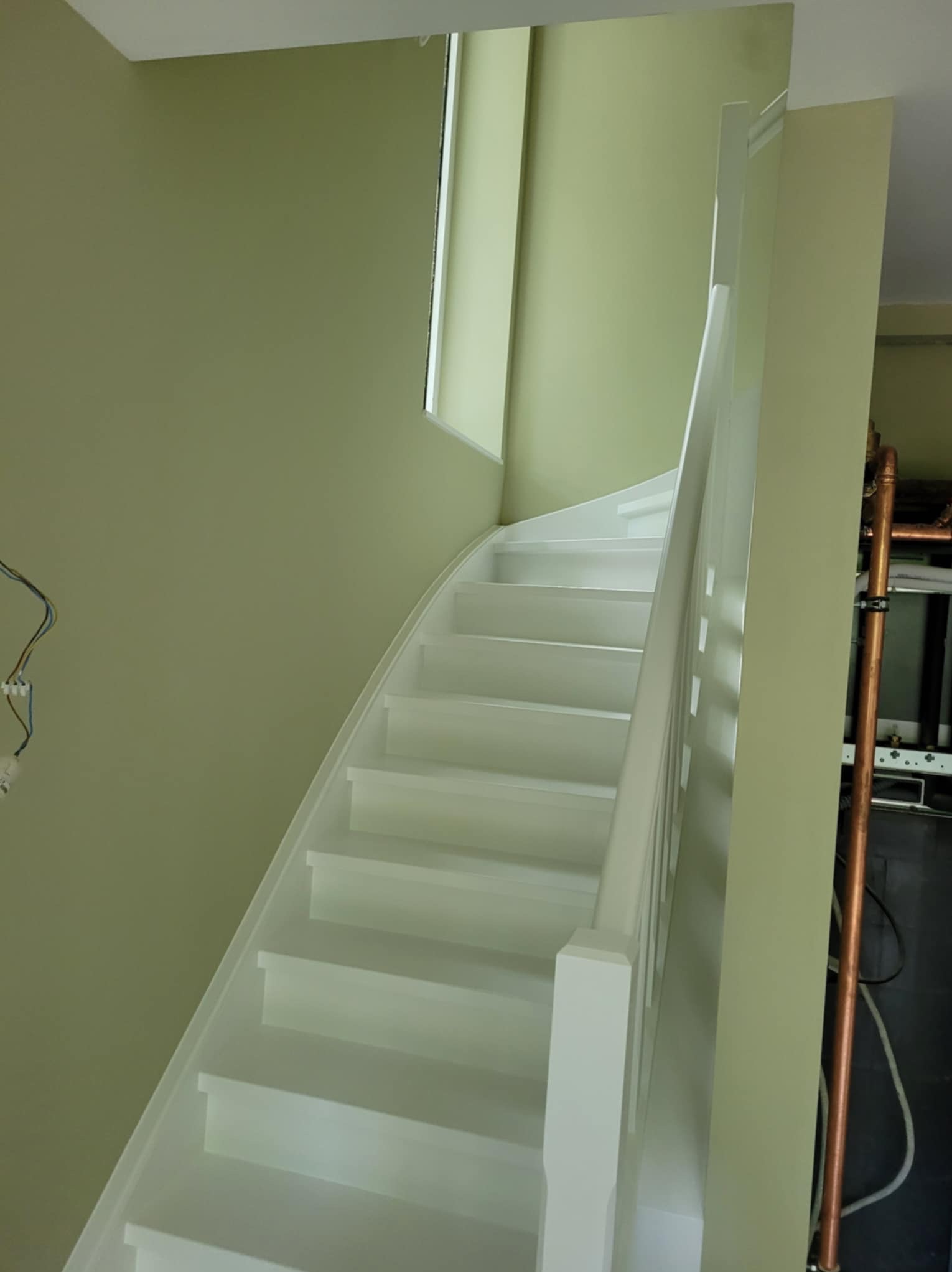 Białe, pomalowane schody kręte na tle oliwkowej ściany z widocznym fragmentem okna i instalacją hydrauliczną w tle
