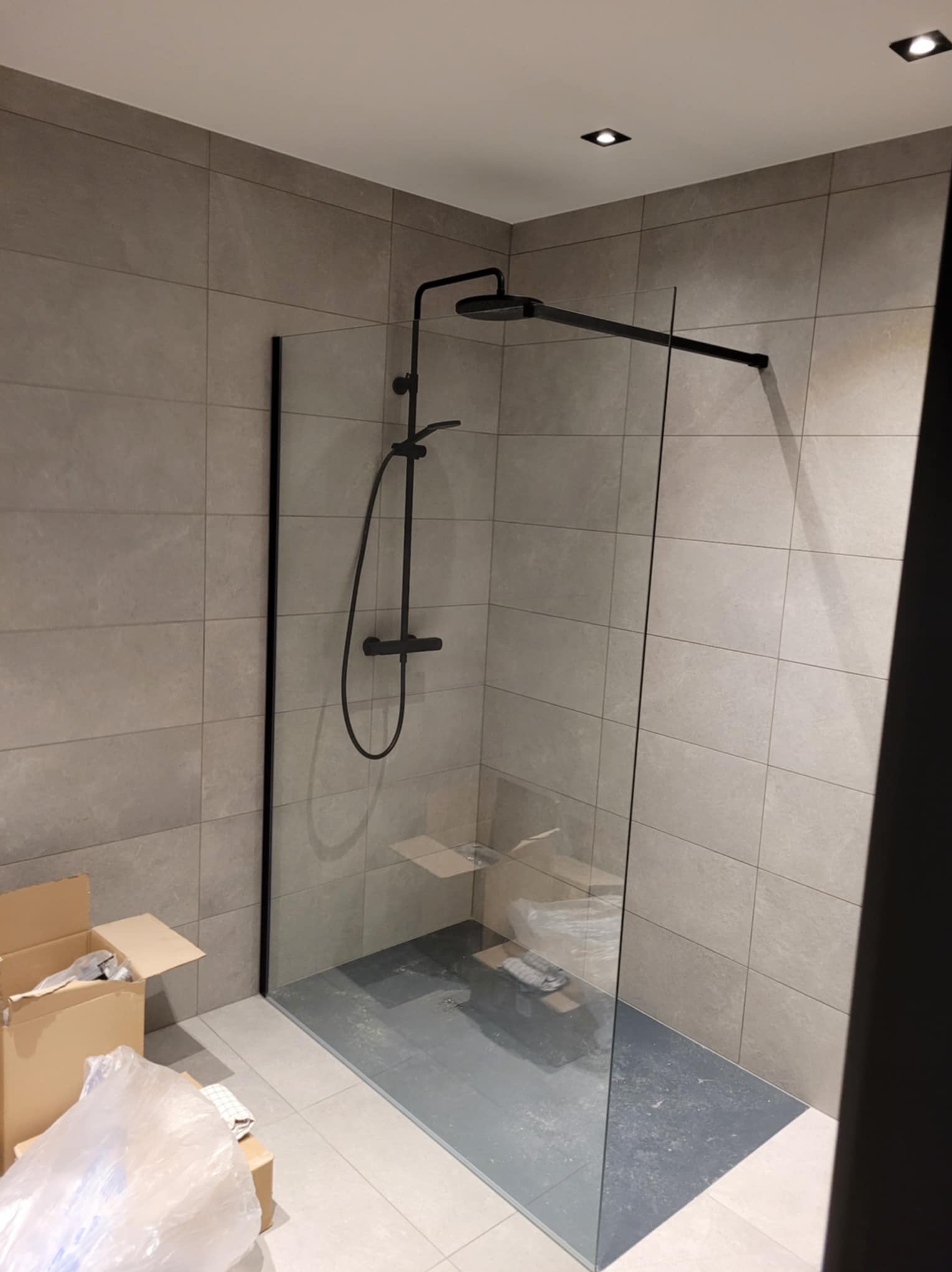 Nowoczesna kabina prysznicowa typu walk-in z czarną armaturą na tle szarych płytek, widoczne kartony i folia ochronna sugerujące prace wykończeniowe.