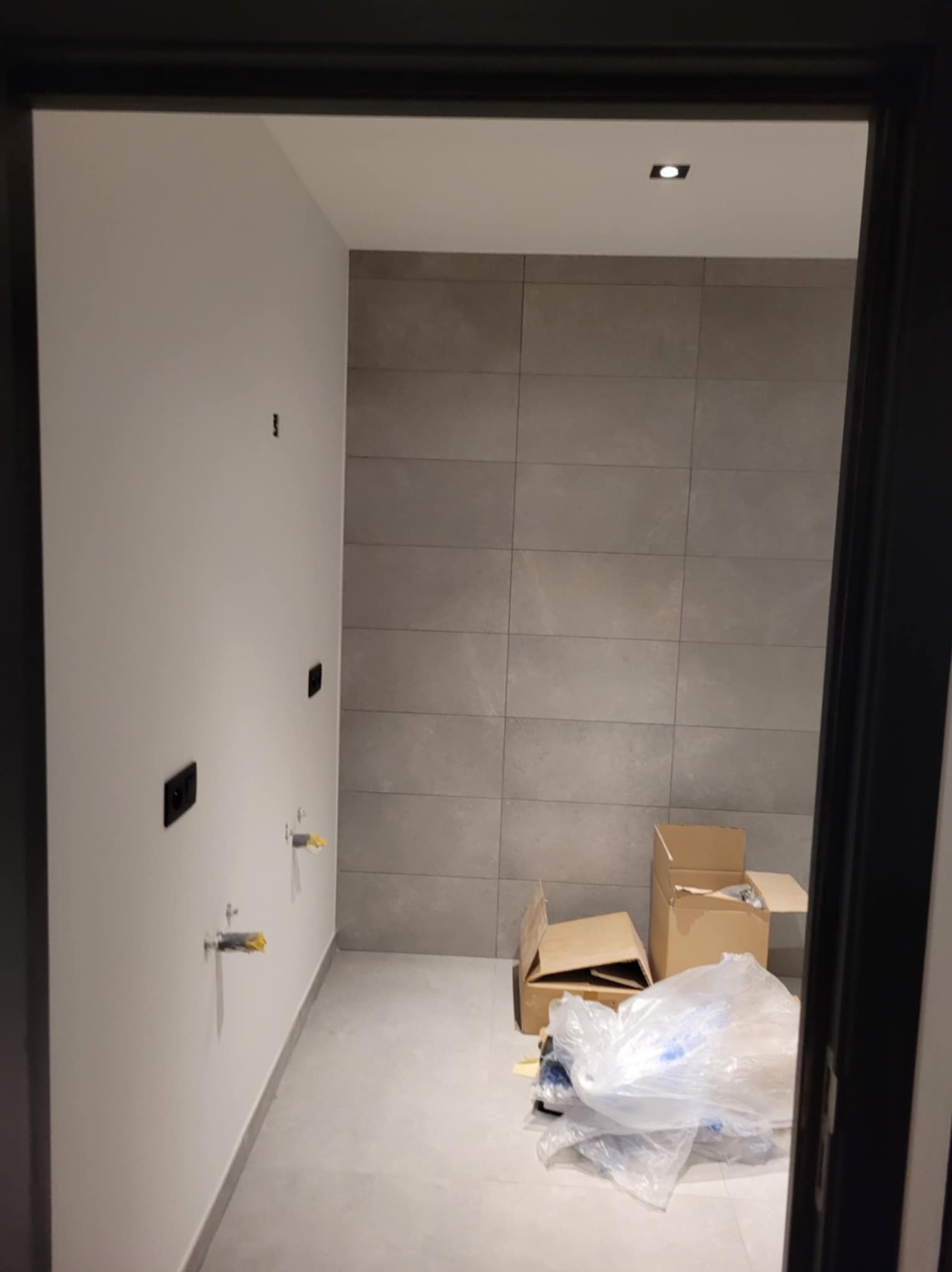 Łazienka w trakcie remontu: szare płytki na ścianie, widoczne wystające rury z żółtymi zabezpieczeniami, niepodłączone gniazdka elektryczne, kartony i folia na podłodze, widok z korytarza przez...