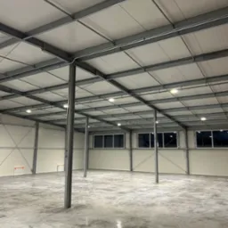 Wnętrze hali przemysłowej z widoczną konstrukcją stalową, oświetleniem LED i betonową posadzką, z fragmentami instalacji elektrycznej poprowadzonej wzdłuż belek stropowych.