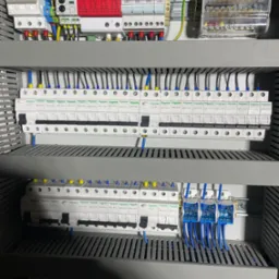 Rozdzielnica elektryczna z zamontowanymi bezpiecznikami i przewodami w kolorach niebieskim, żółtym i czarnym, widoczne zabezpieczenia przeciwprzepięciowe oraz listwa zaciskowa.