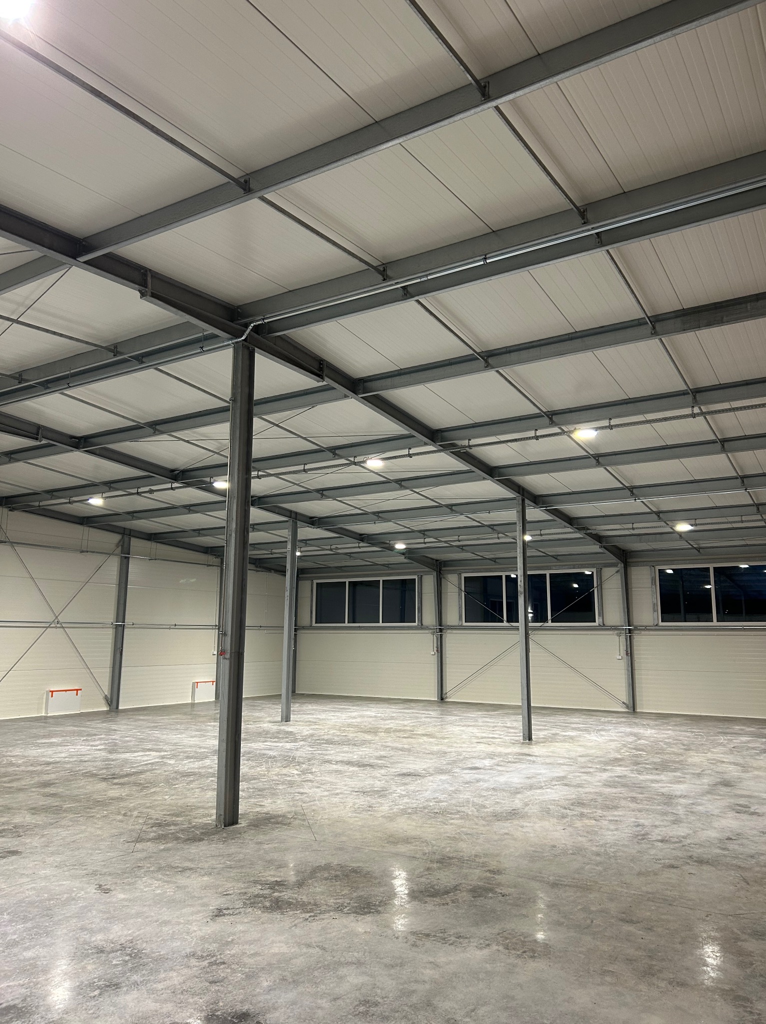 Wnętrze hali przemysłowej z widoczną konstrukcją stalową, oświetleniem LED i betonową posadzką, z fragmentami instalacji elektrycznej poprowadzonej wzdłuż belek stropowych.