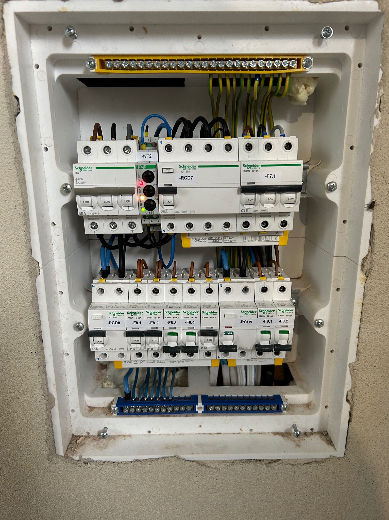 Otwarta skrzynka elektryczna z zamontowanymi bezpiecznikami Schneider Electric, widoczne przewody i listwy zaciskowe, obudowa osadzona w ścianie z widocznymi śladami montażu.