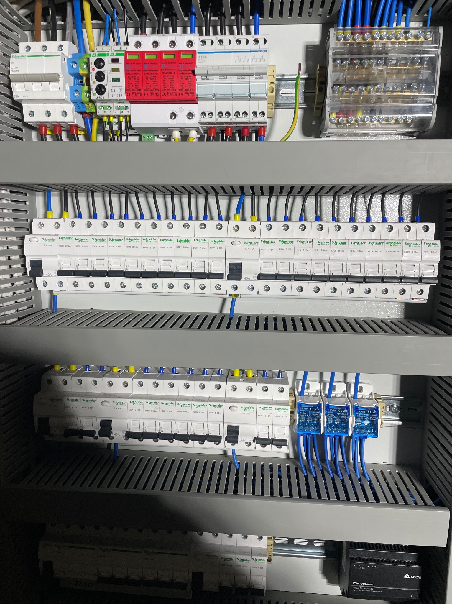 Rozdzielnica elektryczna z zamontowanymi bezpiecznikami i przewodami w kolorach niebieskim, żółtym i czarnym, widoczne zabezpieczenia przeciwprzepięciowe oraz listwa zaciskowa.