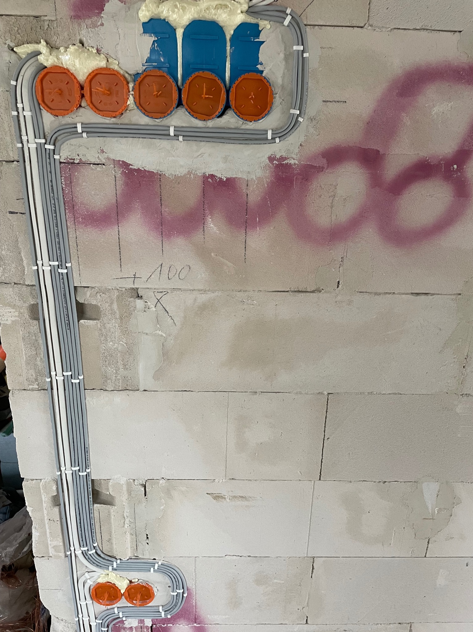 Ściana z betonu komórkowego z widocznymi przewodami elektrycznymi w szarych peszlach, zamocowanymi białymi uchwytami. Pomarańczowe puszki instalacyjne zalane pianką montażową. Widoczne ślady...