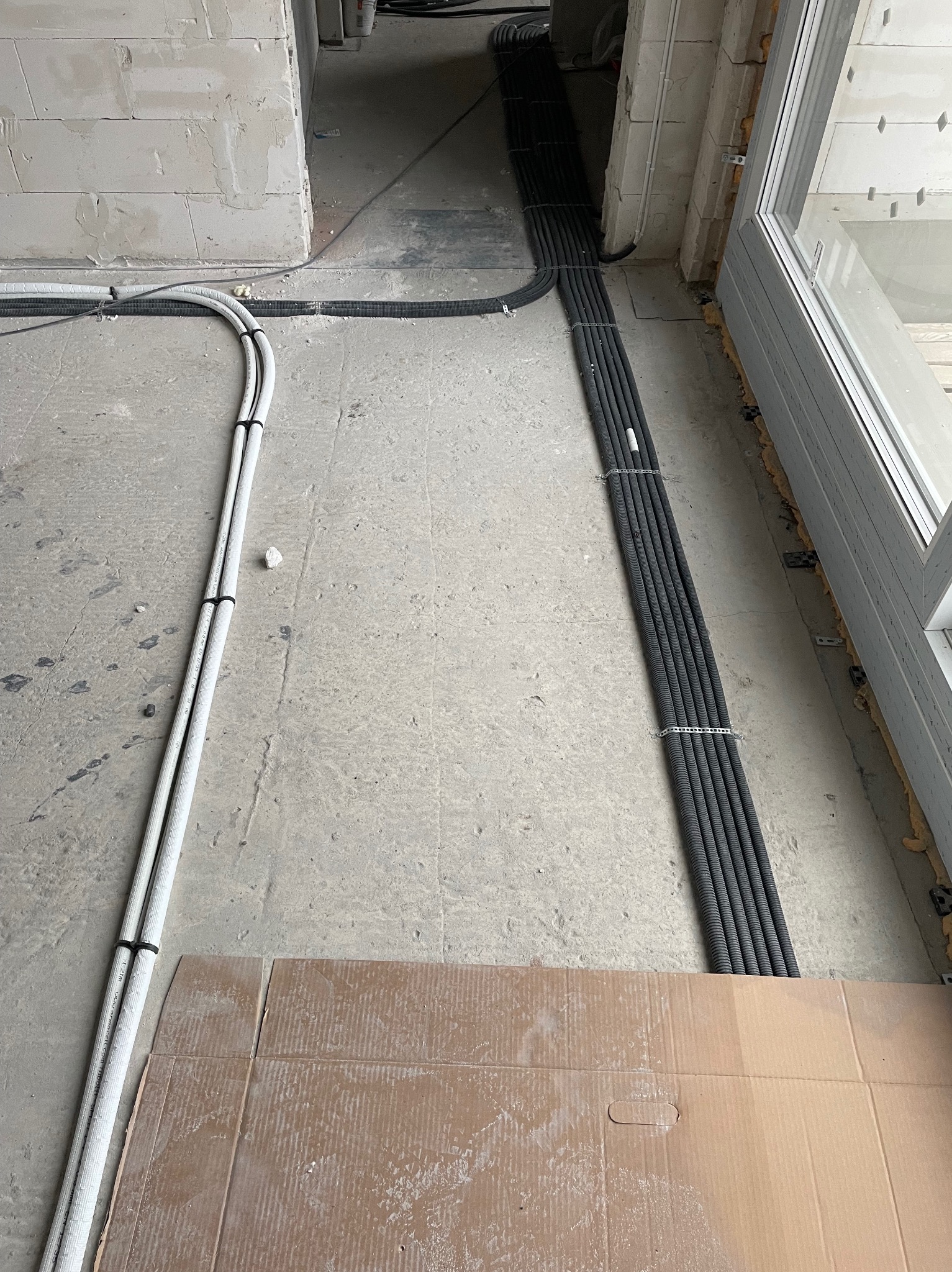 Układanie przewodów elektrycznych w rurach karbowanych, białych i czarnych, na surowym betonie w trakcie prac wykończeniowych w budynku, widoczne fragmenty ściany z bloczków i okna.