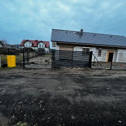 Kompleksowe ogrodzenia - Nowoczesna, czarna brama przesuwna z poziomymi panelami, wkomponowana w ogrodzenie z siatki na tle domu w trakcie budowy, pochmurne niebo.