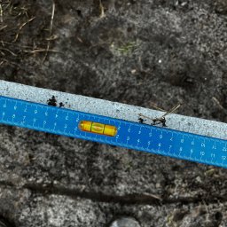 KORSAR Grzegorz Korczyński - Poziomica i miarka na metalowym profilu, prawdopodobnie podczas montażu ogrodzenia. Widoczny fragment buta. Tło to ziemia z resztkami roślin.