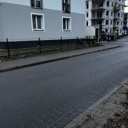 KORSAR Grzegorz Korczyński - Nowoczesny budynek z szarą elewacją i oknami PCV, ogrodzony niskim, czarnym płotem. W tle budowa z dźwigiem. Ujęcie z poziomu chodnika z kostki.