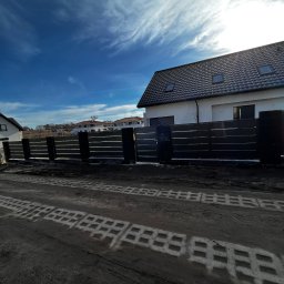 KORSAR Grzegorz Korczyński - Nowoczesne ogrodzenie panelowe w kolorze antracytowym, z poziomymi przęsłami, na tle domu z ciemną dachówką i błękitnego nieba z chmurami. Widoczne ślady opon na ziemi.
