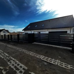 Kompleksowe ogrodzenia - Nowoczesna, pozioma, czarna brama wjazdowa z przesuwnym skrzydłem, widoczna z zewnątrz posesji. Na pierwszym planie ślady po kostce ażurowej.