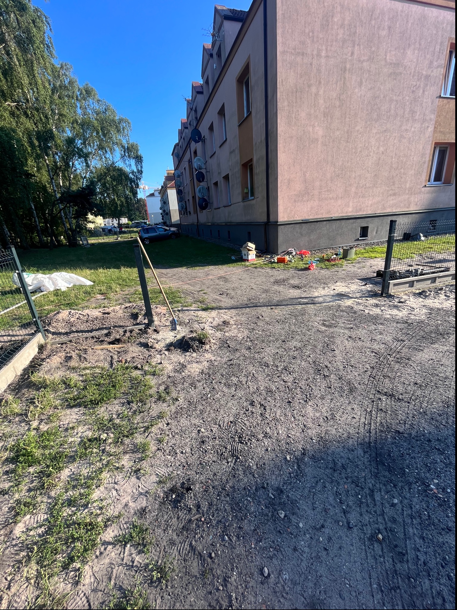 Montaż ogrodzenia panelowego w trakcie: widoczne słupki, ziemia, narzędzie. W tle budynek mieszkalny i zieleń. Kąt ujęcia z dołu podkreśla perspektywę.