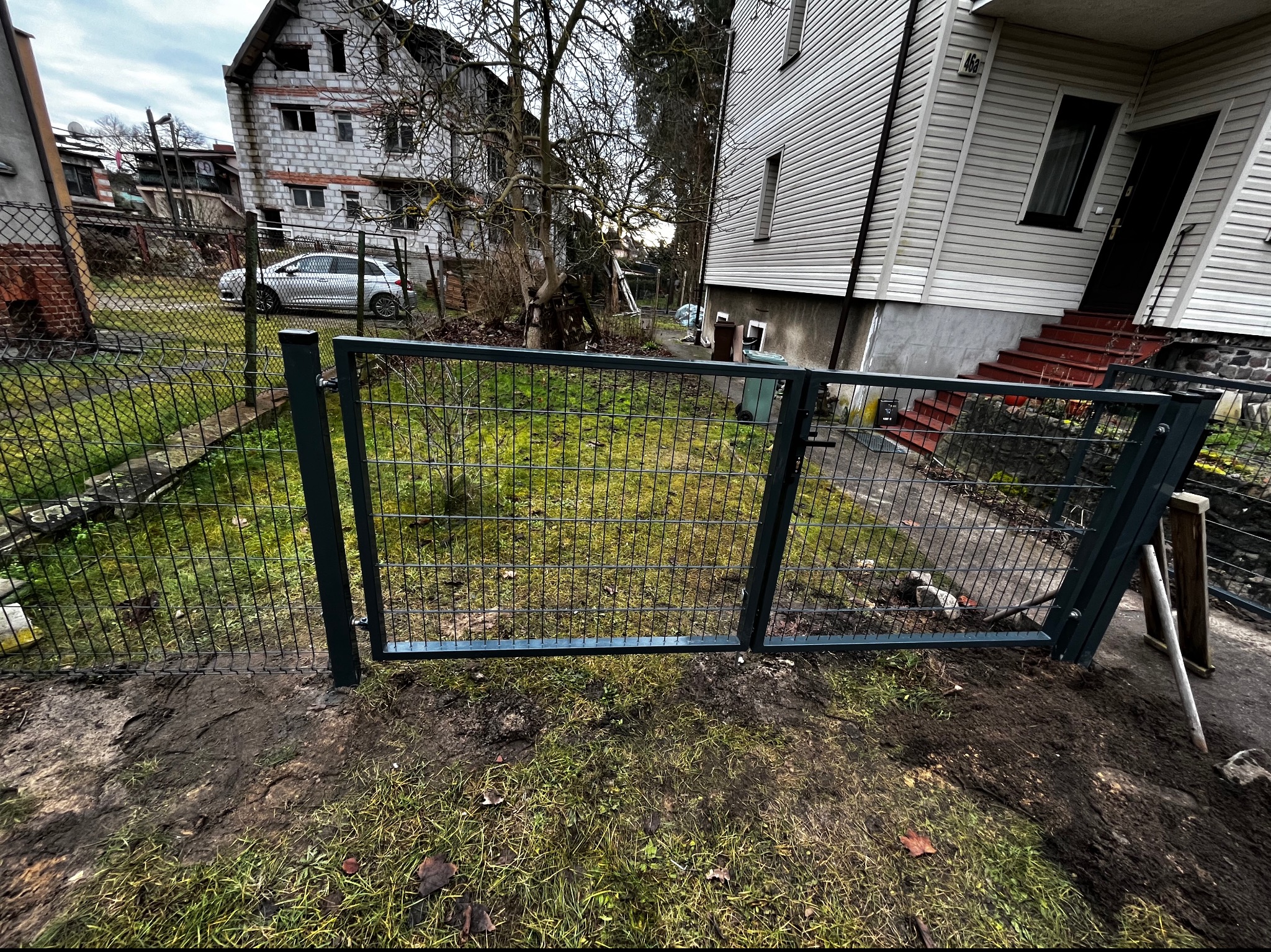 Nowoczesna, metalowa furtka panelowa w kolorze grafitowym, zamontowana na posesji z widocznym trawnikiem i zabudową mieszkalną w tle. Widoczne ślady po montażu.