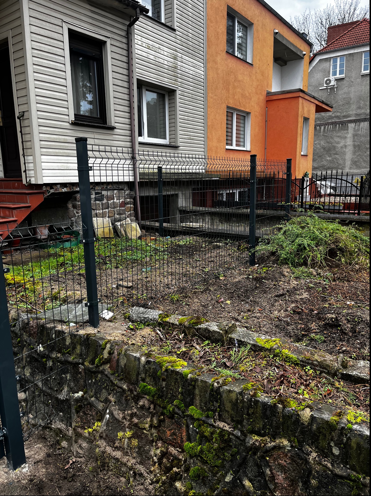 Nowoczesne ogrodzenie panelowe w kolorze grafitowym, oddzielające posesję od ulicy. Widoczne słupki i siatka na tle budynków o różnej architekturze.
