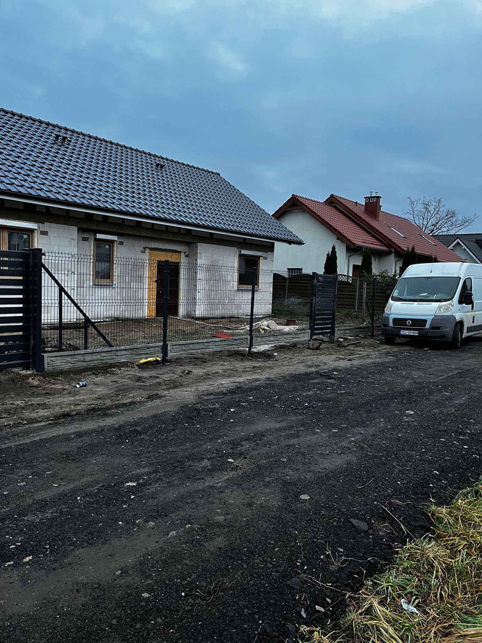 Nowoczesne ogrodzenie panelowe z podmurówką betonową, czarne słupki i przęsła, dom w tle, szara kostka brukowa, biały bus dostawczy. Ujęcie z poziomu gruntu.