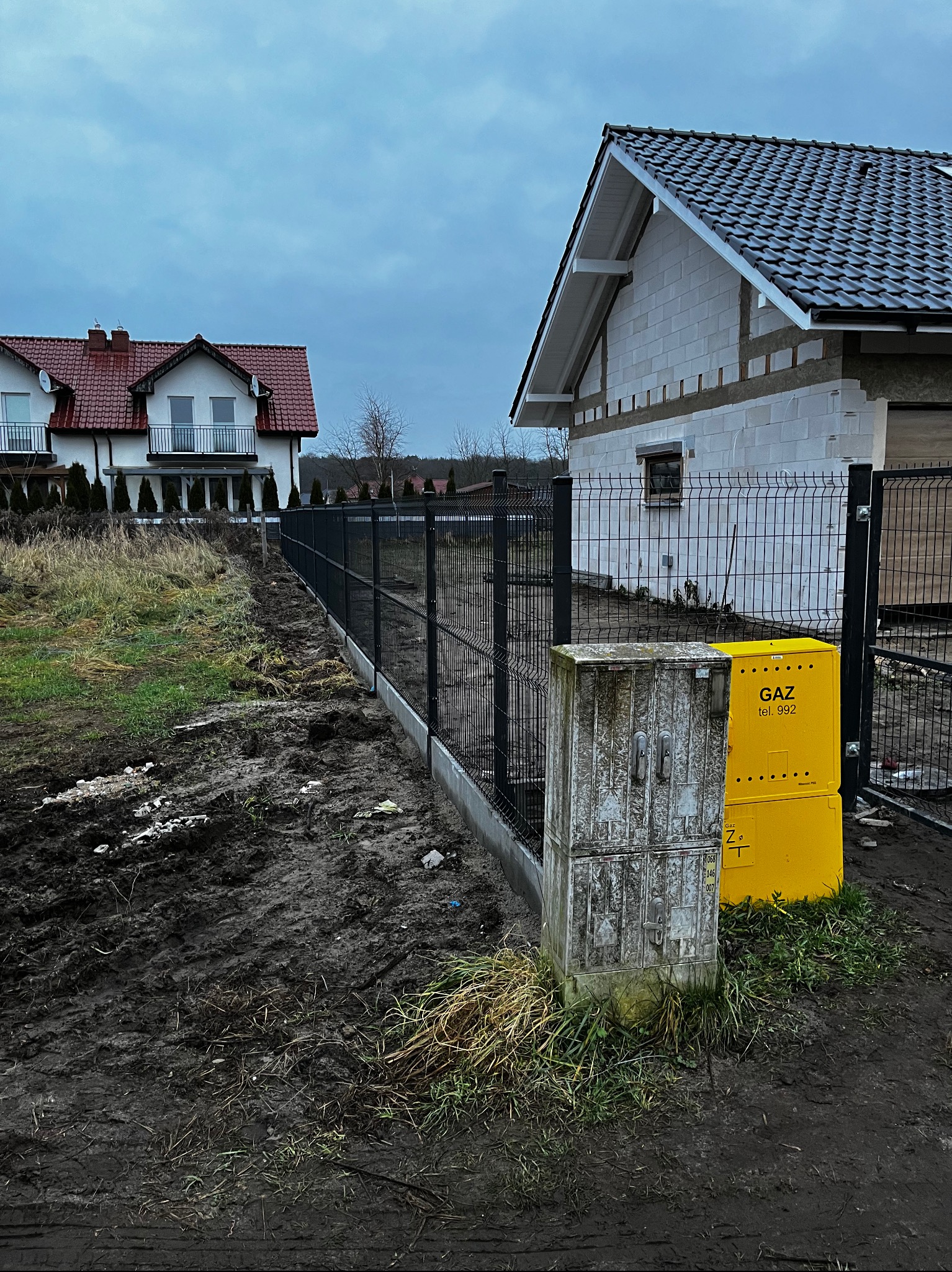 Czarny panelowy płot na betonowej podmurówce oddziela posesje. W tle domy w różnej fazie budowy i skrzynka gazowa. Pochmurny dzień.