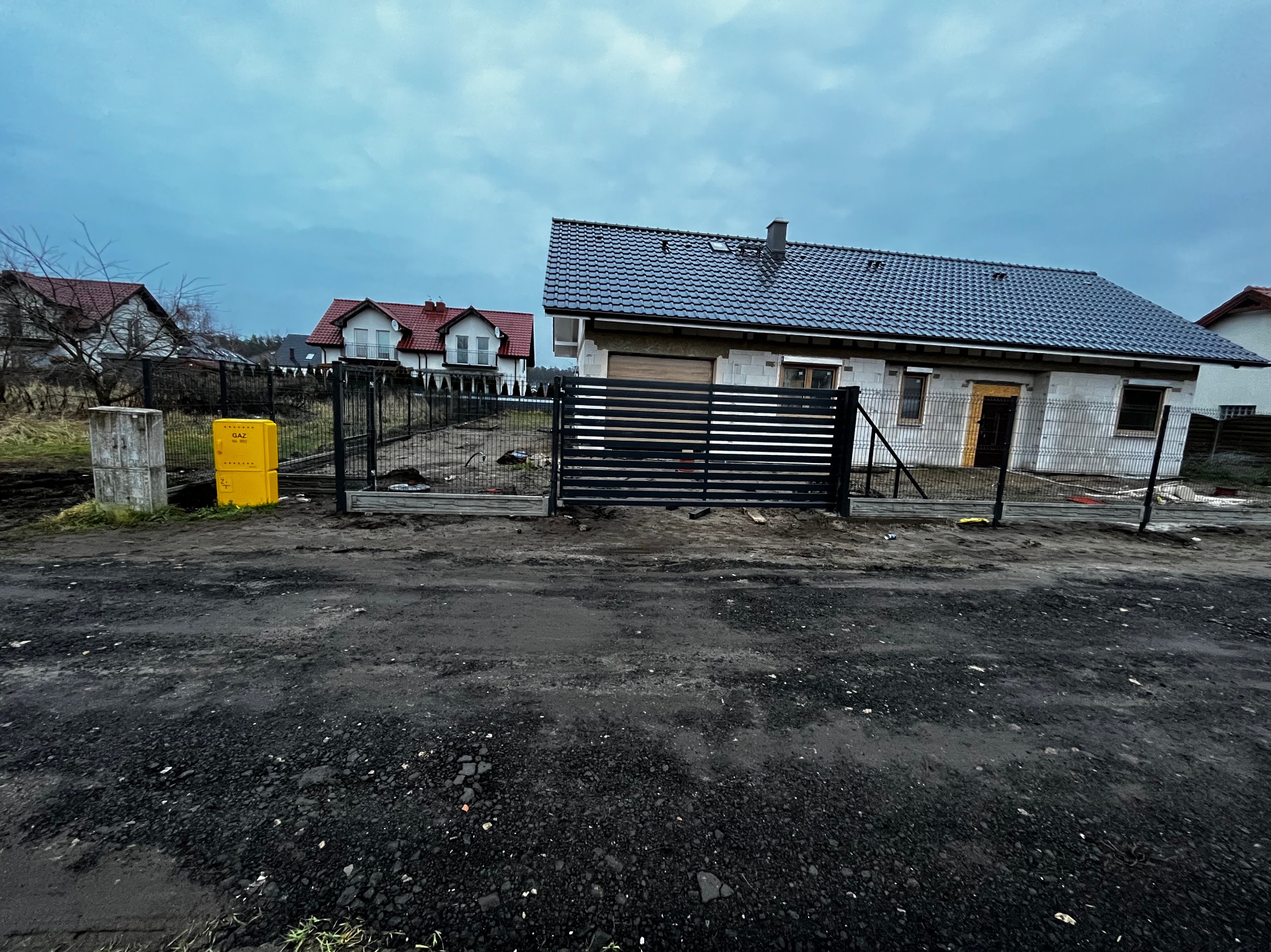 Nowoczesna, czarna brama przesuwna z poziomymi panelami, wkomponowana w ogrodzenie z siatki na tle domu w trakcie budowy, pochmurne niebo.