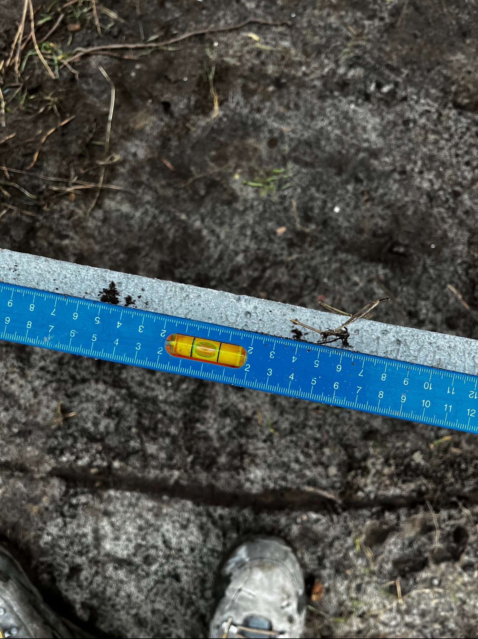 Poziomica i miarka na metalowym profilu, prawdopodobnie podczas montażu ogrodzenia. Widoczny fragment buta. Tło to ziemia z resztkami roślin.